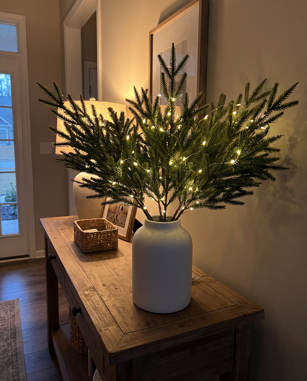 Styled illustration: 10 Glowbough pine stems with a warm light strand in a white vase on an entryway console at dusk