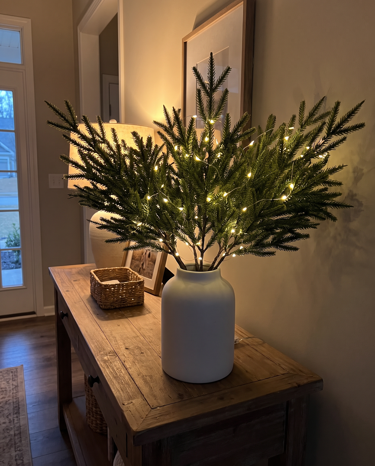 Styled illustration: 10 Glowbough pine stems with a warm light strand in a white vase on an entryway console at dusk