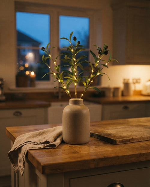 Styled illustration: three lit faux olive stems glowing in a stone ceramic vase on an oak kitchen island at dusk
